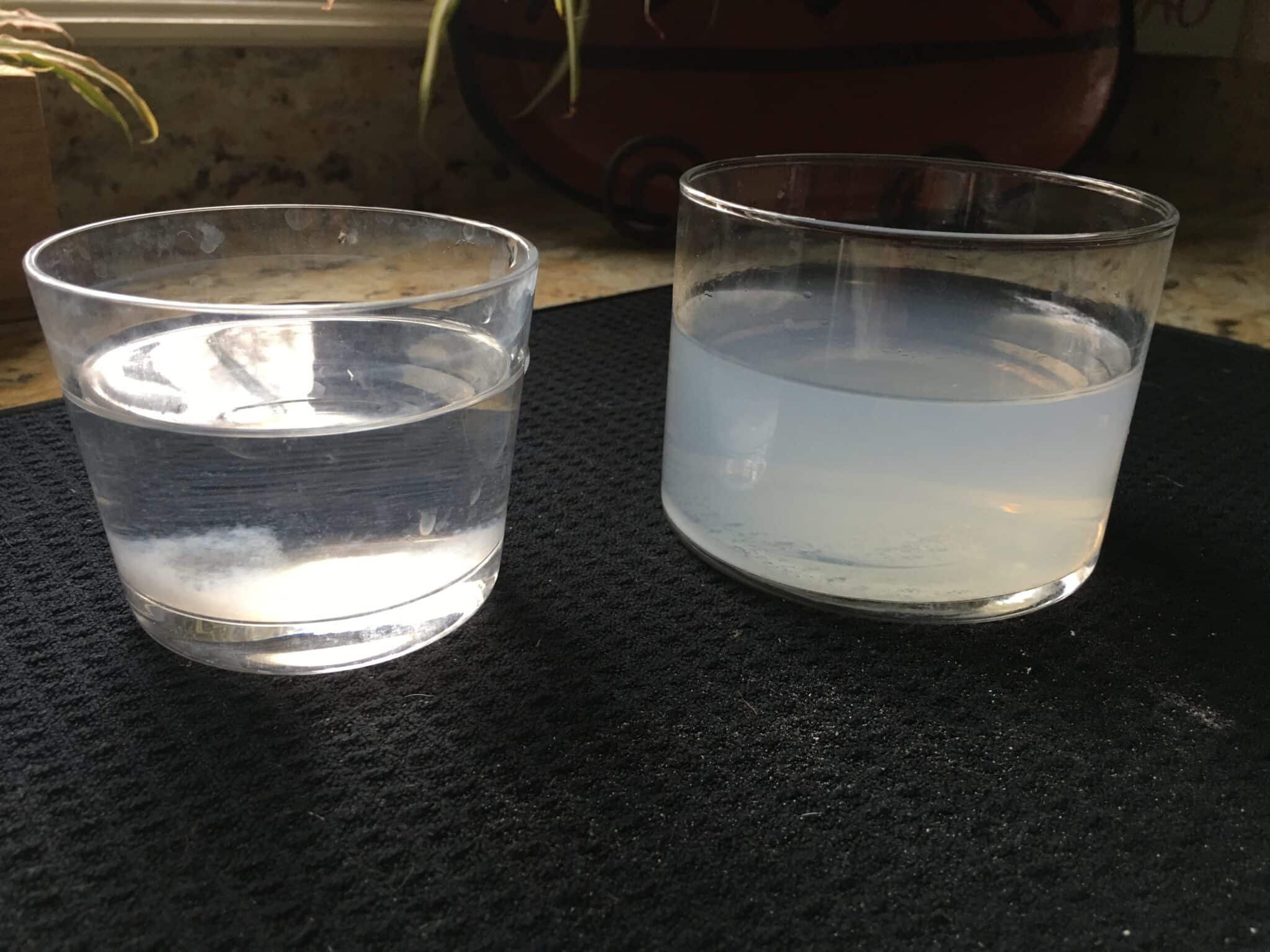Two clear glasses sit on a black mat. The glass on the left contains clear water, while the glass on the right contains cloudy, white water. Both are placed near a kitchen countertop with faint sunlight.