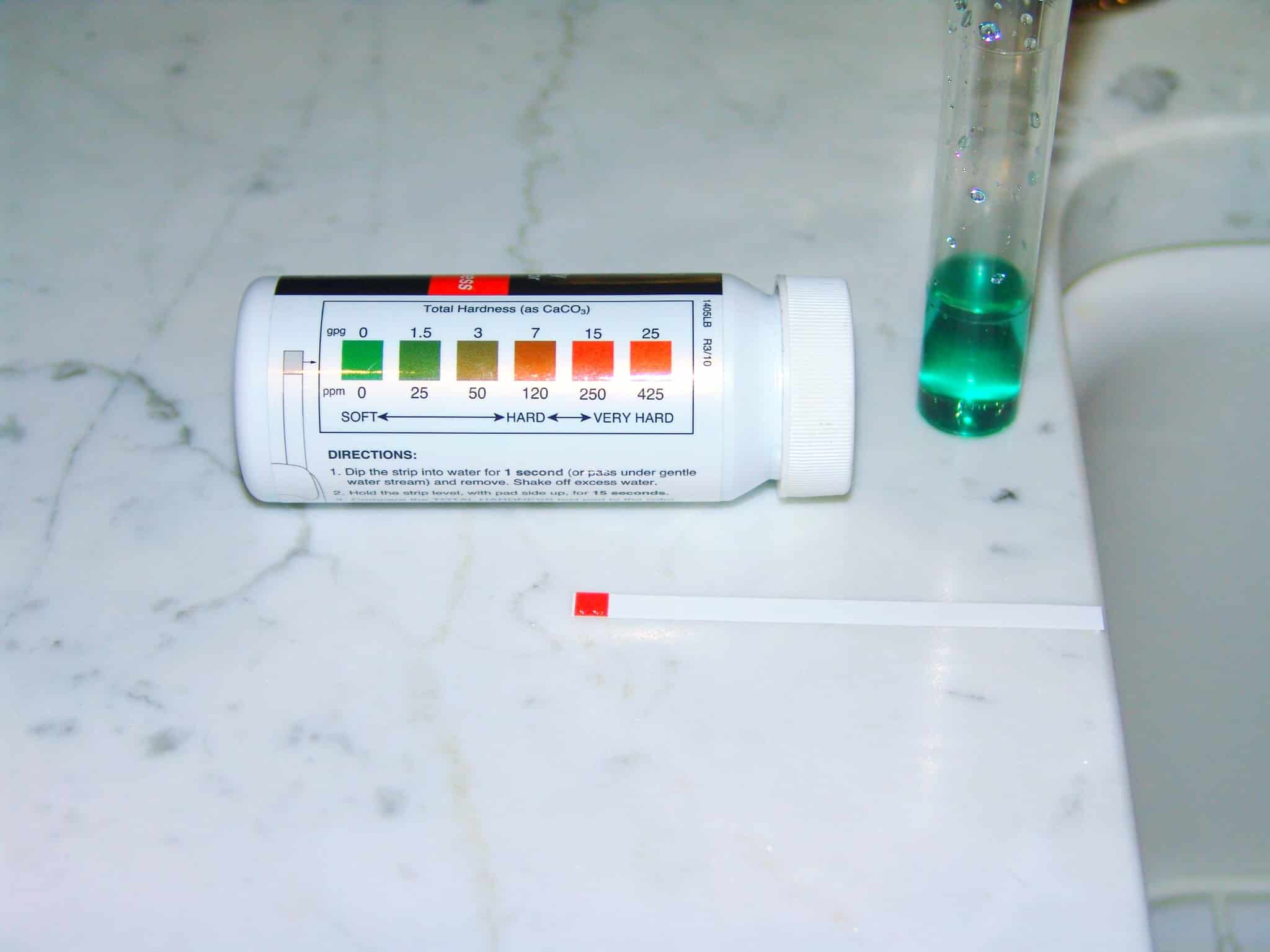 A water hardness test kit with a labeled container, a test strip, and a vial of green liquid on a marble counter. The label shows a color chart indicating softness to hardness levels.