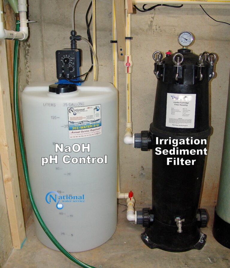 A large white tank labeled NaOH pH Control sits next to a tall black cylinder labeled Irrigation Sediment Filter in a utility area, with pipes, gauges, and control equipment attached.