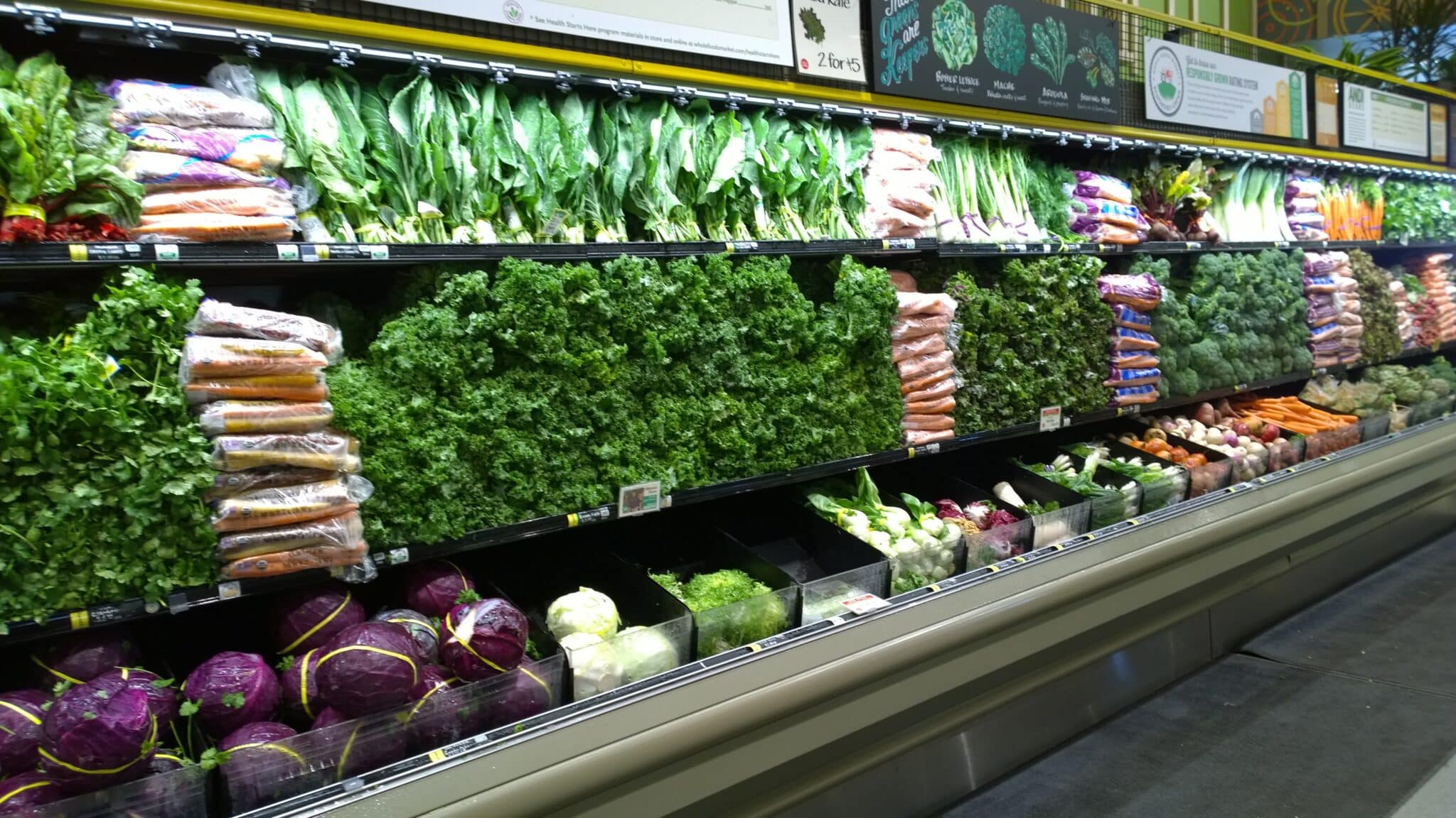 A grocery store produce section with shelves neatly stocked with fresh vegetables, including carrots, lettuce, kale, purple cabbage, celery, and other leafy greens.