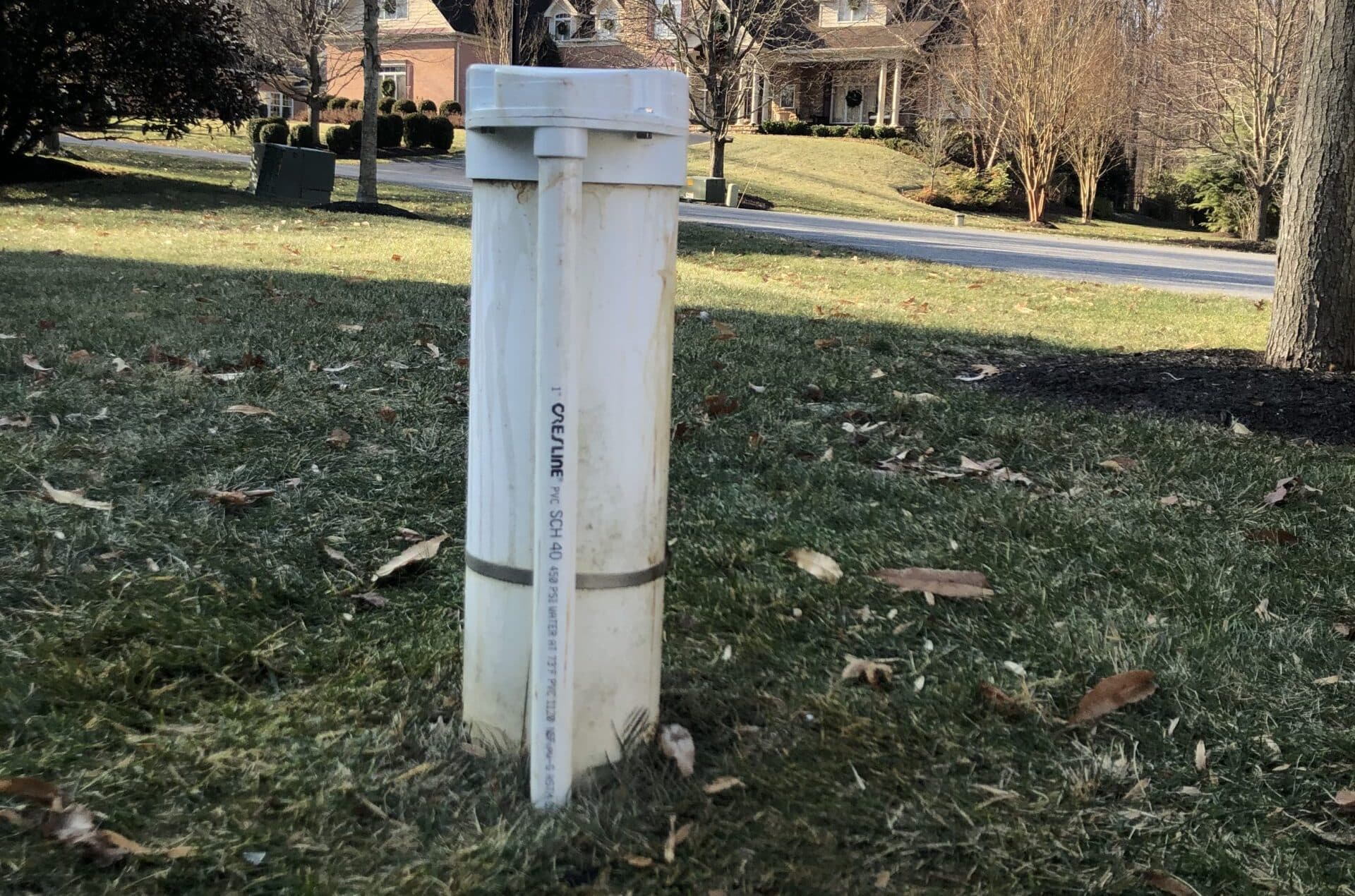 A white PVC well pipe with a capped top stands upright in a grassy yard with scattered leaves, in front of suburban homes and a street.