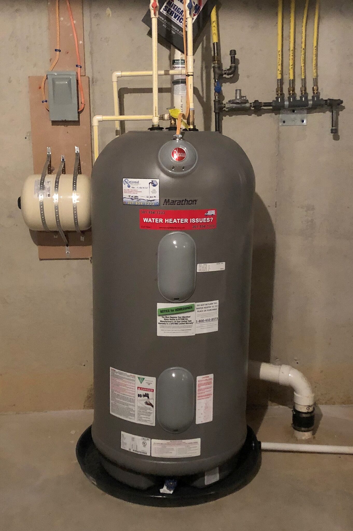 A gray Marathon water heater stands in a basement with pipes connected at the top and sides, labels on the front, and a black drainage pan underneath. Yellow gas lines and a control box are mounted on the concrete wall behind it.