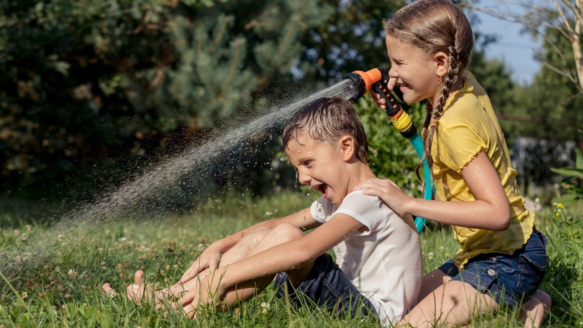 Two children play outside on the grass; a girl sprays a garden hose at a smiling boy, both laughing and enjoying the sunny weather.