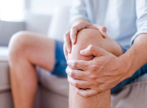 A person wearing shorts sits on a couch, holding their knee with both hands, suggesting discomfort or pain in the knee area, possibly from exposure to acidic water.