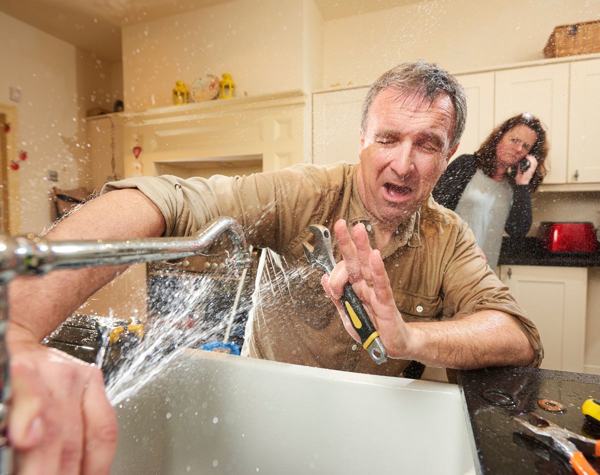 A man struggles to fix a kitchen sink as water sprays everywhere, drenching him. He winces and holds up a wrench. In the background, a woman stands by the counter, likely calling about whole home water treatment systems.