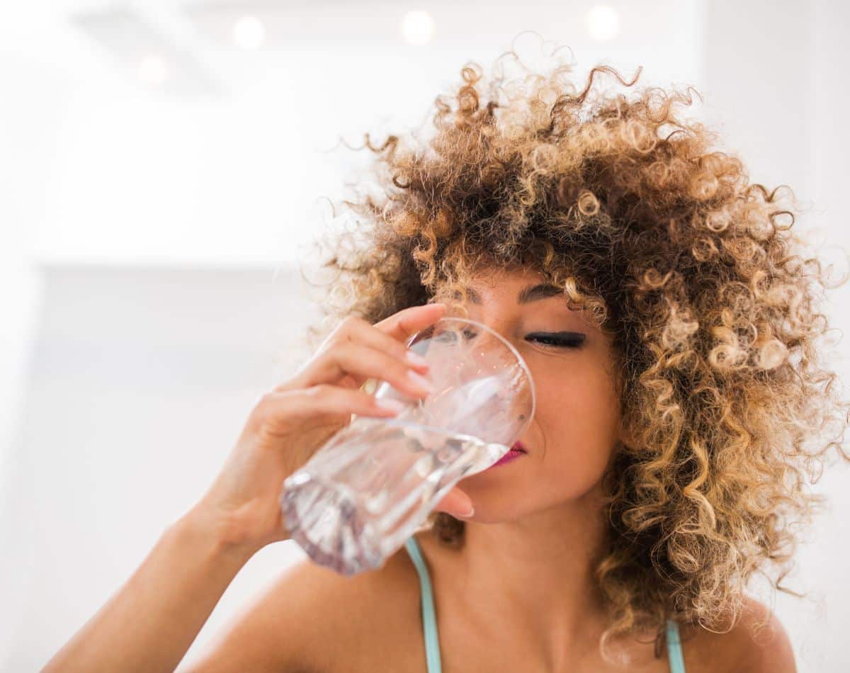 A woman with curly hair drinks a glass of water indoors, her eyes closed as she enjoys a refreshing sip—thanks to whole home water treatment systems. The background is bright and softly blurred.