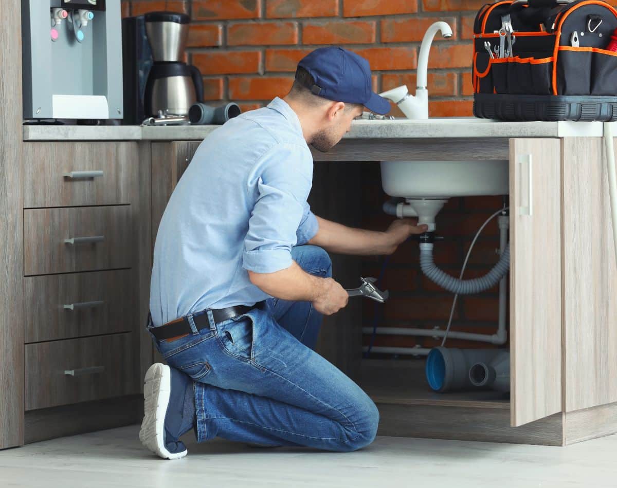 A plumber wearing a blue cap and shirt kneels in front of an open kitchen cabinet, fixing the pipes under a sink with a wrench—a tool bag on the counter nearby suggests he may be installing whole home water treatment systems.