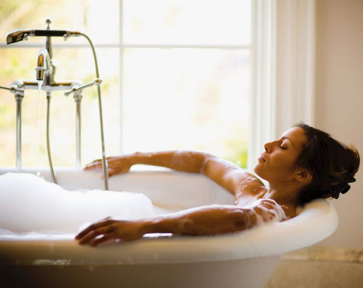 A woman relaxes in a bubble-filled bathtub near a window, resting her head back with eyes closed and arms draped on the tub’s edge, enjoying a peaceful moment thanks to her reliable water heater.