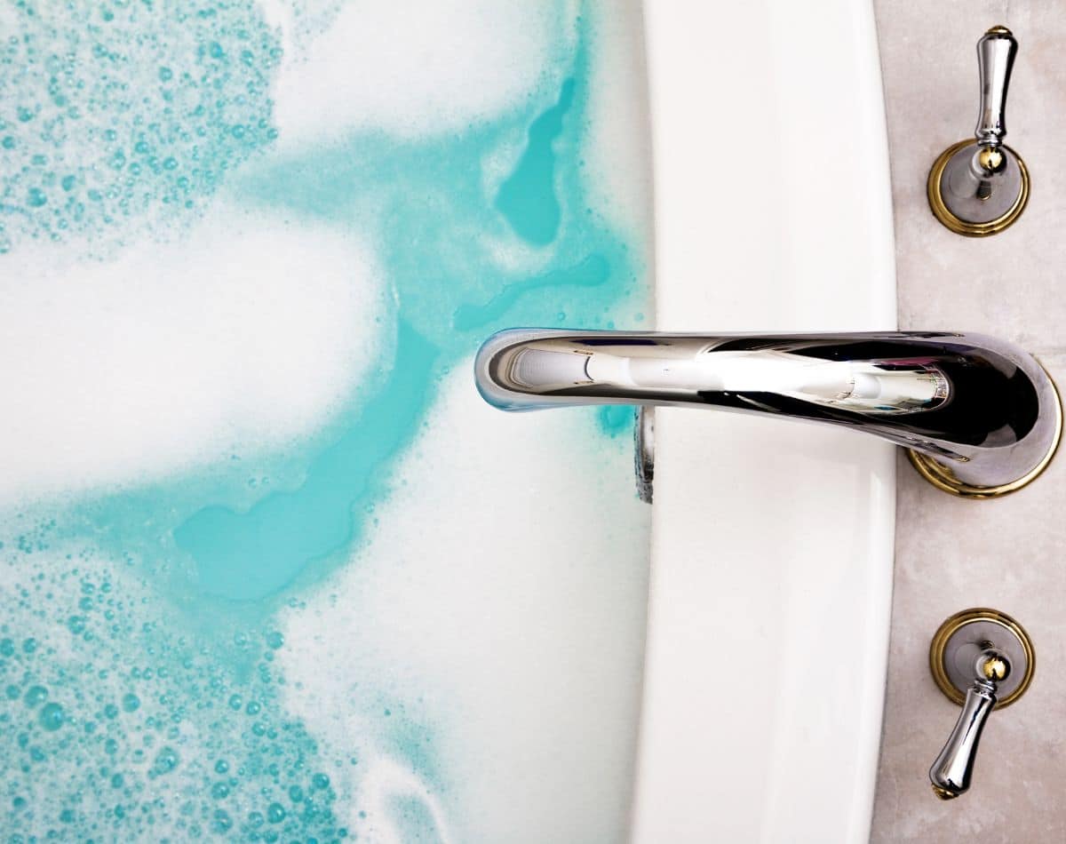 Close-up of a bathtub filled with light blue water and white bubbles, thanks to a reliable water heater. A shiny silver faucet and two matching handles are visible on the right side of the image.