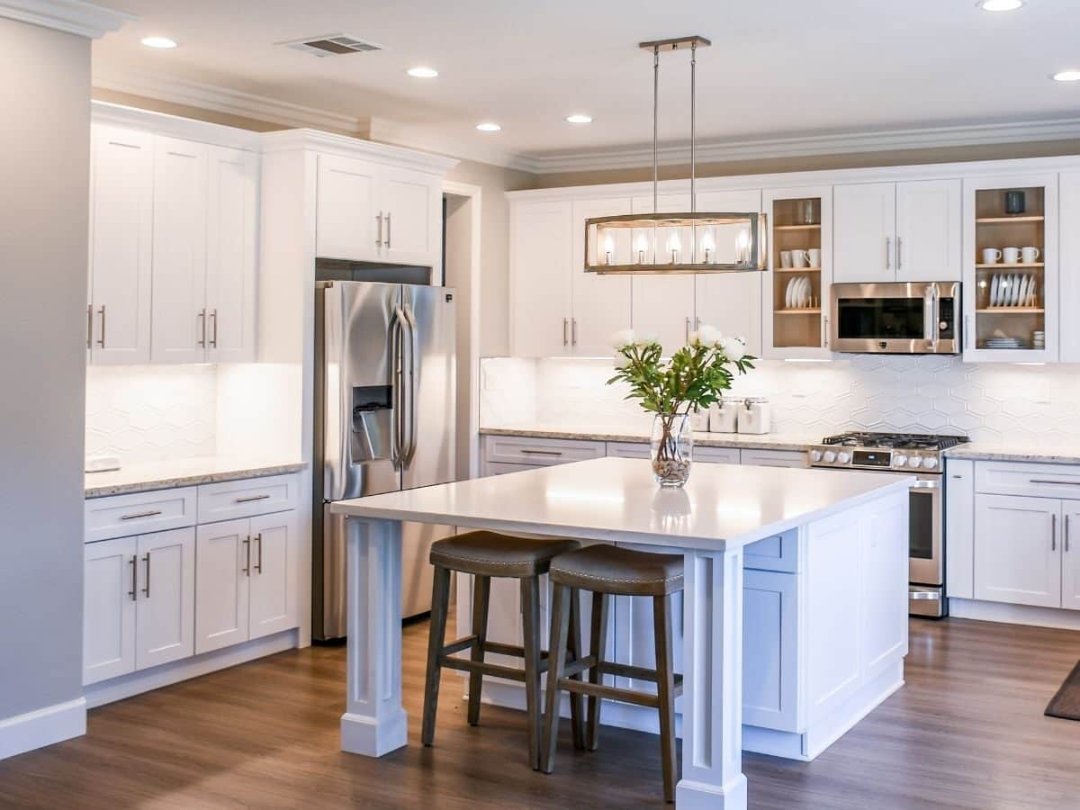 Modern kitchen with white cabinets, stainless steel appliances, a center island with two stools, a vase with flowers, and wood flooring. Bright recessed lighting and a rectangular fixture enhance the space—ideal for when you need a plumber for your kitchen.