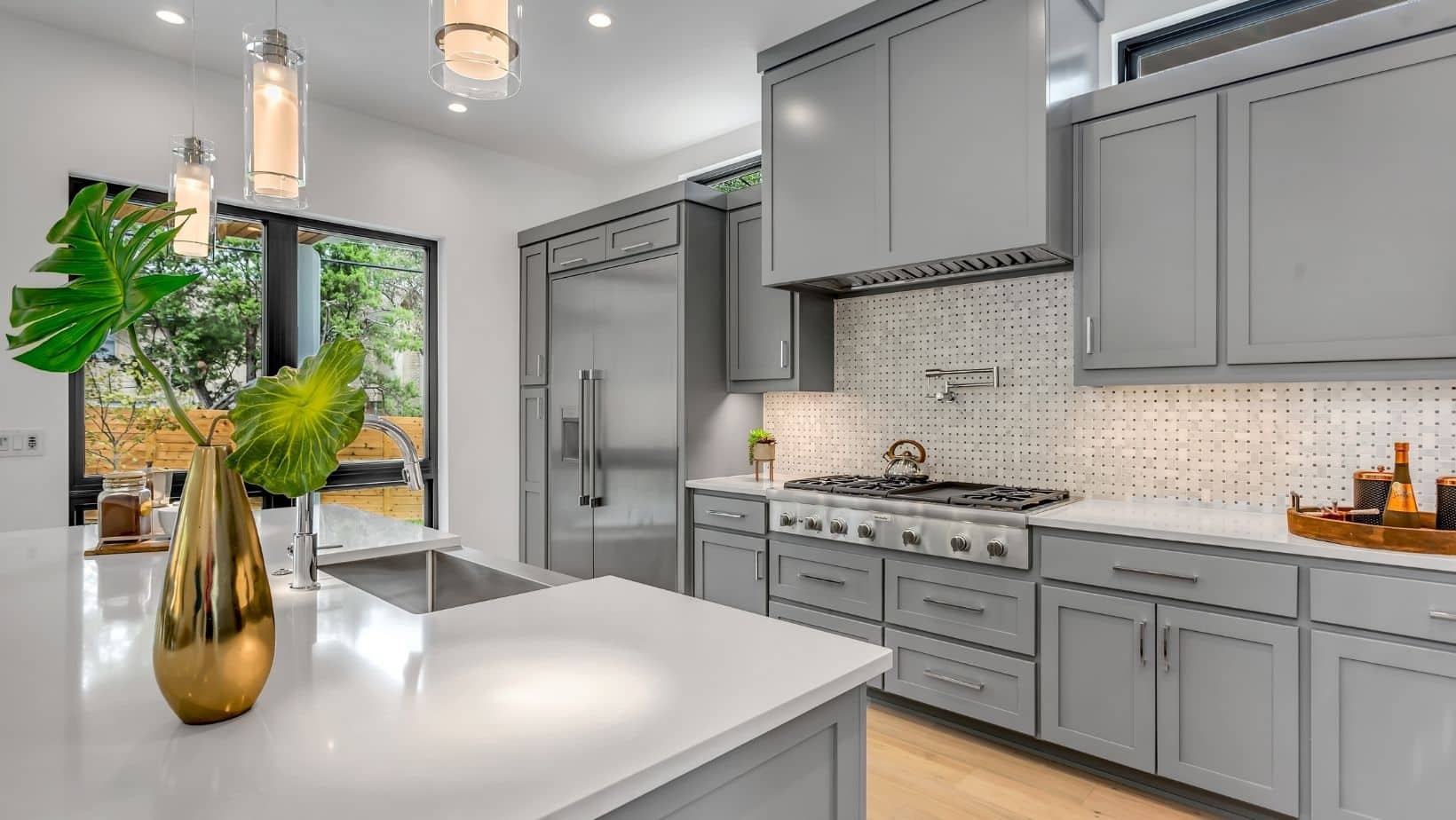 Modern kitchen with light gray cabinets, stainless steel appliances, a gas stove, and white countertops. For installations or repairs, consider a reliable plumber for your kitchen. Large windows fill the space with natural light.