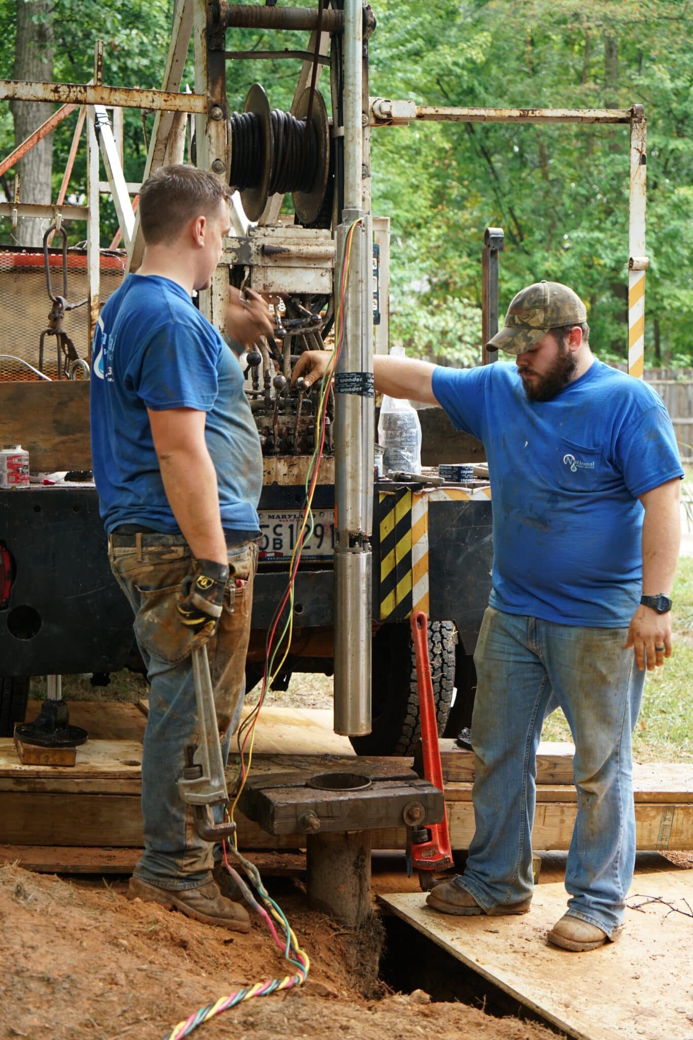 Two men in blue shirts work with machinery on a platform outdoors. One man holds a metal pipe while the other gestures, both surrounded by tools and cables, with trees visible in the background.