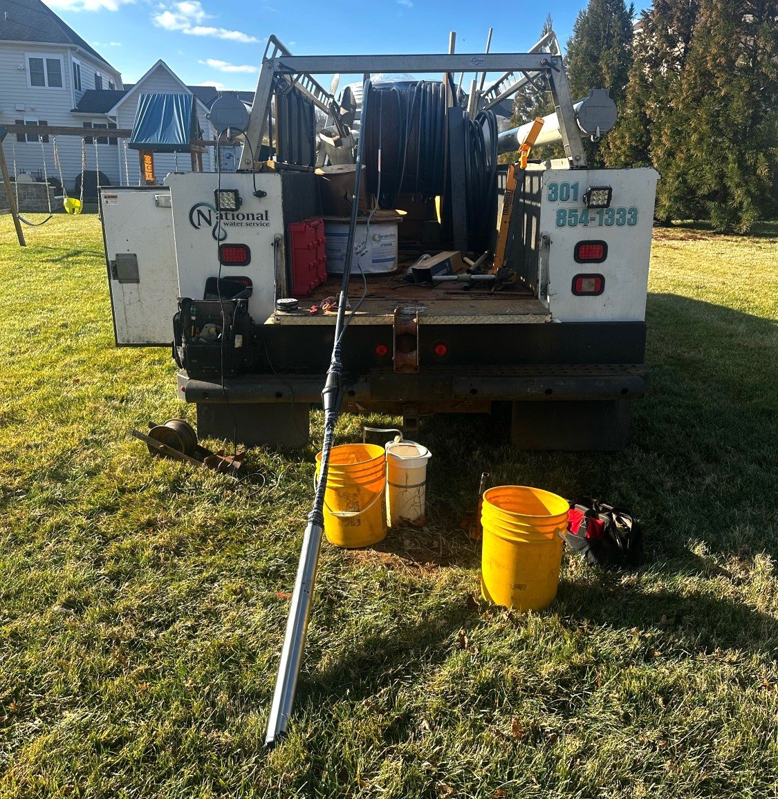 A work truck with equipment and hoses parked on grass, with three yellow buckets and tools laid out behind it. Houses and trees are visible in the background on a sunny day.