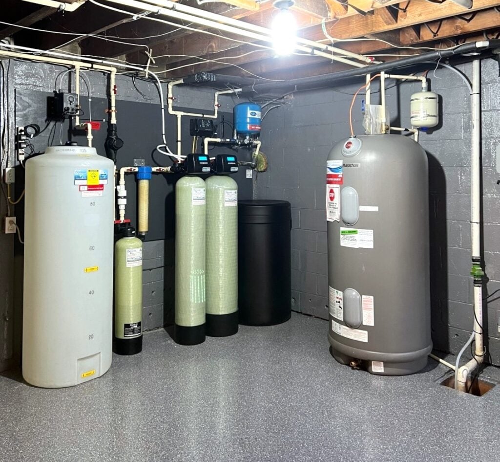 A basement utility area with large whole home water treatment tanks, filtration systems, a water heater, pipes, and electrical wiring mounted on painted cinderblock walls and an exposed wood beam ceiling. The floor is clean and gray.