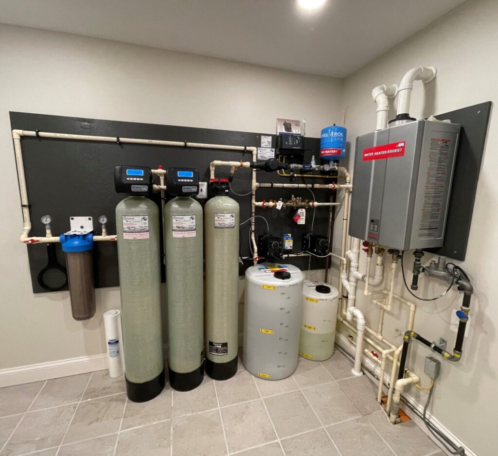 A whole home water treatment system installed in a tiled room, featuring large filter tanks, pipes, control panels, pressure tanks, and a wall-mounted hot water heater unit.