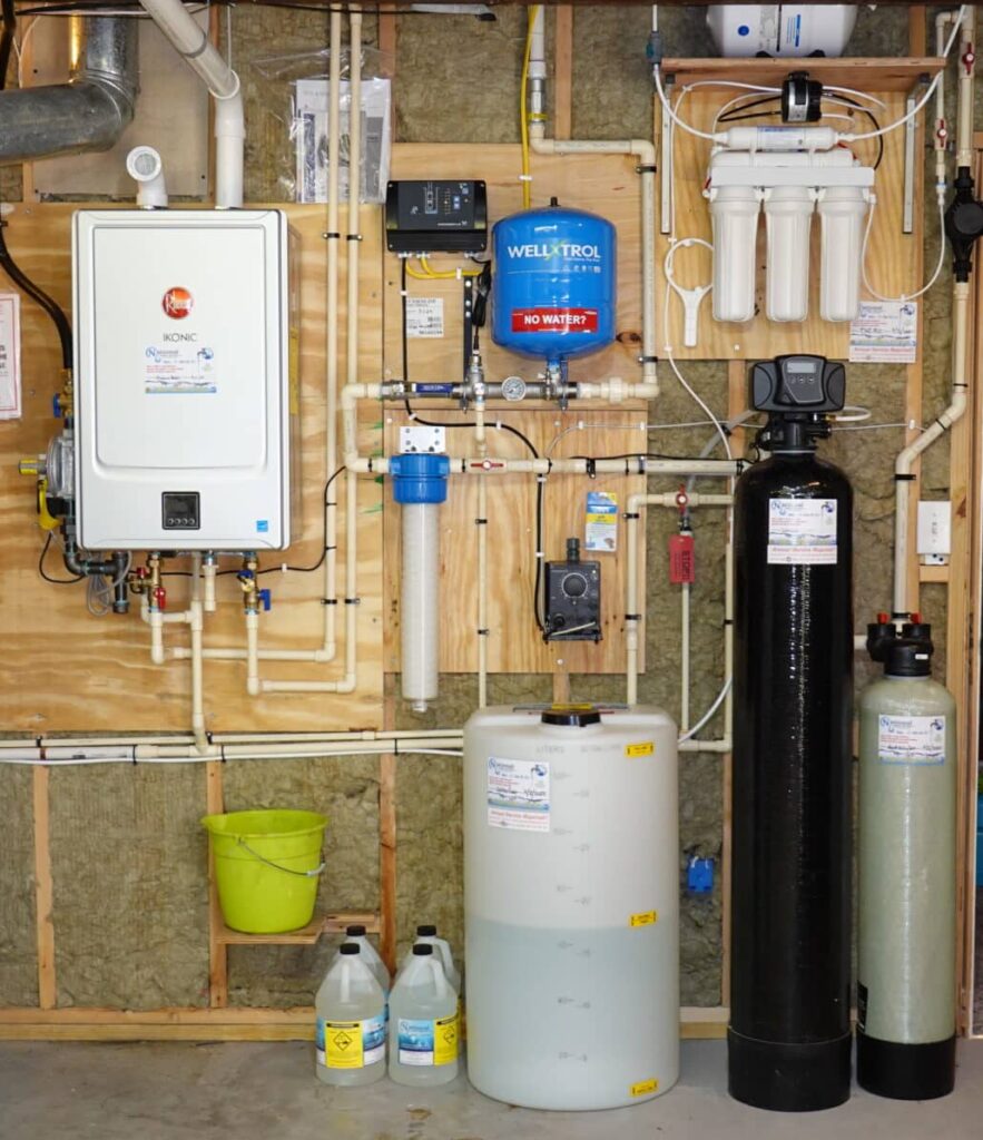 A residential Home Water Treatment system is mounted on a basement wall, featuring tanks, filters, pipes, gauges, and bottles of solution, all organized against a wooden and insulation backdrop.