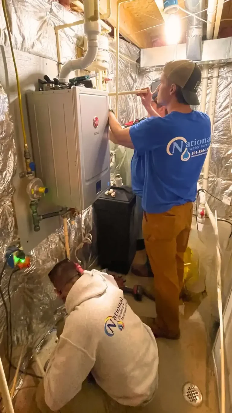 Two workers in National Water Service shirts install or repair plumbing equipment in a basement with metallic insulated walls. One works on pipes, while the other crouches near the floor, both focused on water treatment tasks.