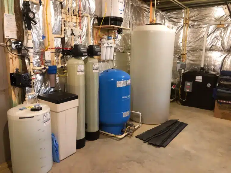 A Maryland Water Treatment system featuring large tanks, a blue pressure tank, water filtration units, and pipes is installed against a wall with reflective insulation. The concrete floor remains clear and clean.