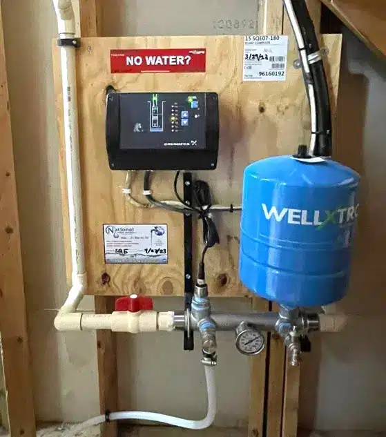 A residential Maryland Well Pump control system mounted on a wooden board, featuring a digital display, pressure gauge, blue Well-X-Trol tank, pipes, and a red valve; a NO WATER? sign is displayed above the monitor.