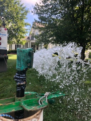Water gushes forcefully from a green outdoor well pump, spraying droplets in the air. A house and trees are visible in the blurred background on a sunny day, highlighting the effectiveness of quality well pump installation.