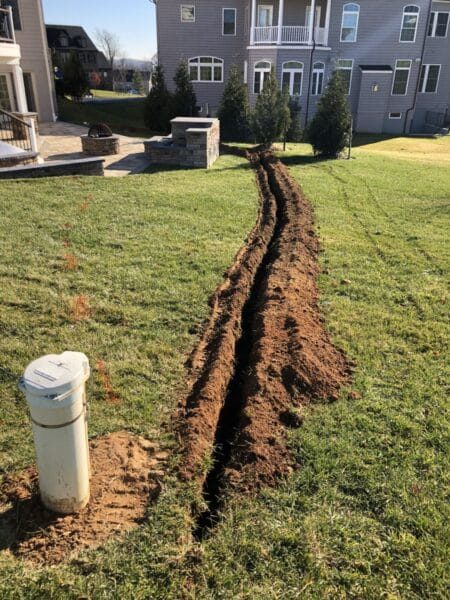 A long, narrow trench is dug through a grassy backyard leading from a white utility pipe toward a modern house, with soil piled on both sides—suggesting ongoing Maryland Well Pump repair work.