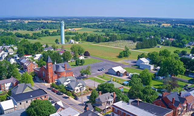 Aerial view of a small town with houses, a red brick building, water treatment Carroll County MD facility, a water tower, parking lots, green fields, and trees under a clear blue sky.