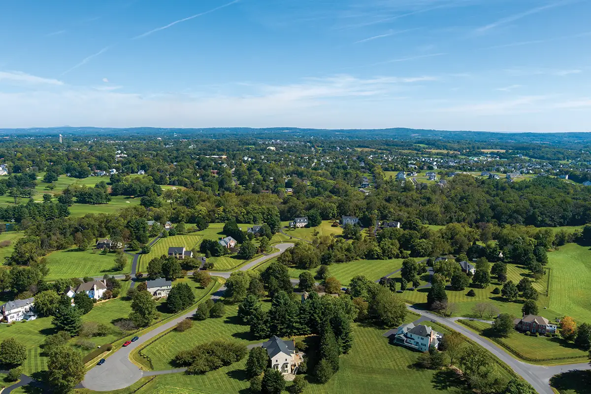Aerial view of a suburban area in Loudoun County, VA, with scattered houses, green lawns, winding roads, and rolling hills under a blue sky—showcasing a community benefiting from advanced water treatment Loudoun County services.