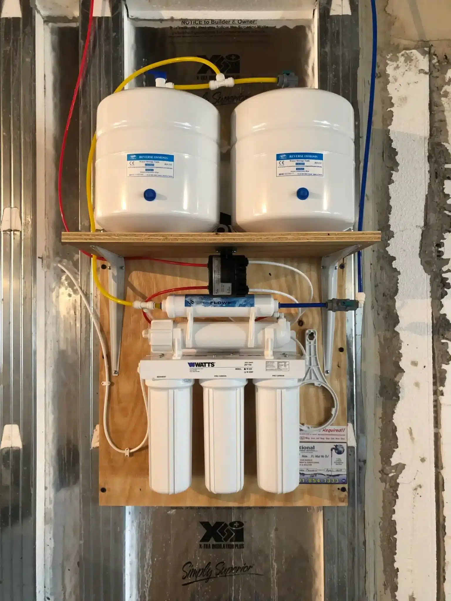 A wall-mounted reverse osmosis water filtration system with three vertical filter cartridges below two white water storage tanks, connected by colored tubing, installed on a wooden board in a partially finished room.