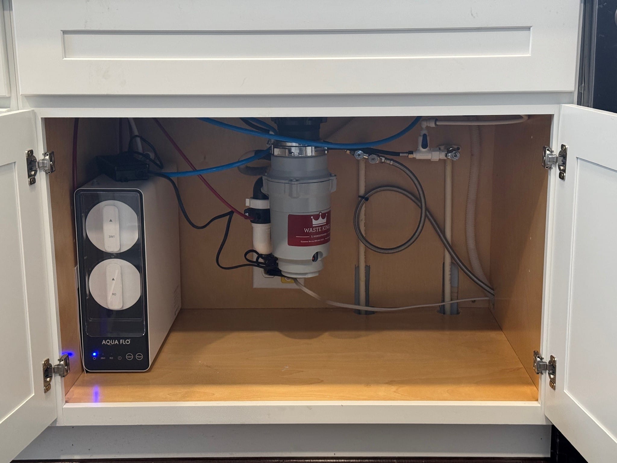Open cabinet showing an under-sink setup with a Reverse Osmosis water filtration system on the left, electrical outlets, and a garbage disposal unit mounted to the sink above, with various pipes and wiring visible. Open cabinet showing an under-sink setup with a Reverse Osmosis water filtration system on the left, electrical outlets, and a garbage disposal unit mounted to the sink above, with various pipes and wiring visible.