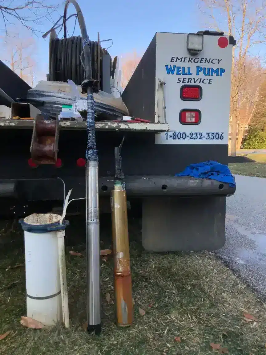 Two submersible well pumps, one clean and one rusted, stand upright on grass beside a white utility truck labeled “Emergency Well Pump Service” with a phone number on the back. Tools and equipment are on the truck.
