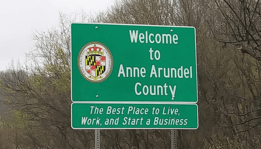 Green roadside sign reading Welcome to Anne Arundel County with the county seal. Below, a smaller sign says, The Best Place to Live, Work, and Start a Business. Trees and water treatment Anne Arundel County facilities are visible in the background.