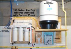 Reverse osmosis water filtration system and 14.4-gallon storage tank mounted on a wall, with pipes, labeled controls, and shelves holding a wrench and manuals; text reads “200 Gallon Per Day Reverse Osmosis with Booster Pump.”. Reverse osmosis water filtration system and 14.4-gallon storage tank mounted on a wall, with pipes, labeled controls, and shelves holding a wrench and manuals; text reads “200 Gallon Per Day Reverse Osmosis with Booster Pump.”.