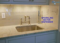 Close-up of a kitchen sink with a gold faucet and a modern metallic sparkling water dispenser, set against a beige subway tile backsplash and granite countertop. Blue text reads SPARKLING WATER DISPENSER. Close-up of a kitchen sink with a gold faucet and a modern metallic sparkling water dispenser, set against a beige subway tile backsplash and granite countertop. Blue text reads SPARKLING WATER DISPENSER.