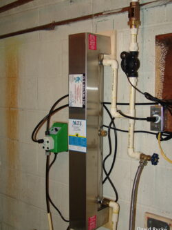 A UV water purification system mounted on a basement wall, connected to plastic pipes and valves, with a green control box and several labels on the unit. A UV water purification system mounted on a basement wall, connected to plastic pipes and valves, with a green control box and several labels on the unit.