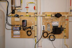 A water filtration and pump control system is mounted on a wall with pipes, valves, wires, control boxes, and labeled components attached to wooden boards. A water filtration and pump control system is mounted on a wall with pipes, valves, wires, control boxes, and labeled components attached to wooden boards.