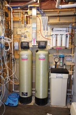 A home water filtration system in a basement, featuring two large vertical tanks, a filtration unit with gauges and tubes, and various wires and pipes attached to a wooden wall. A blue salt bag is on the floor. A home water filtration system in a basement, featuring two large vertical tanks, a filtration unit with gauges and tubes, and various wires and pipes attached to a wooden wall. A blue salt bag is on the floor.