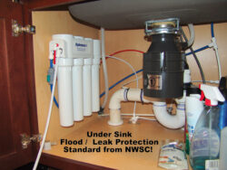 Under a kitchen sink, a water filtration system and garbage disposal are installed among pipes and cleaning supplies. Text reads: Under Sink Flood / Leak Protection Standard from NWSC!. Under a kitchen sink, a water filtration system and garbage disposal are installed among pipes and cleaning supplies. Text reads: Under Sink Flood / Leak Protection Standard from NWSC!.