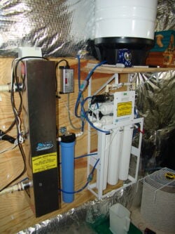 A home water filtration system with multiple filter cartridges, tubes, and control units is mounted on a wooden wall in a basement or utility room with reflective insulation. A blue filter and various labels are visible. A home water filtration system with multiple filter cartridges, tubes, and control units is mounted on a wooden wall in a basement or utility room with reflective insulation. A blue filter and various labels are visible.