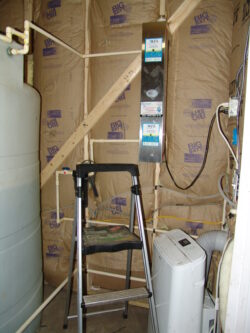 A small utility room with exposed insulation, a step ladder, a white plastic tank, a water filter system mounted on the wall, and a portable air conditioning unit with a vent hose. A small utility room with exposed insulation, a step ladder, a white plastic tank, a water filter system mounted on the wall, and a portable air conditioning unit with a vent hose.