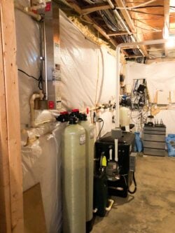 A basement with exposed ceiling beams, various water filtration tanks, pipes, electrical wiring, and equipment lining the walls. The walls are covered with white plastic sheeting, and the floor is unfinished concrete. A basement with exposed ceiling beams, various water filtration tanks, pipes, electrical wiring, and equipment lining the walls. The walls are covered with white plastic sheeting, and the floor is unfinished concrete.