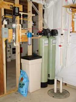 A home water softener system in a basement, featuring two tall green tanks, a brine tank with a Solar Salt bag beside it, piping, and electronic controls mounted on unfinished wooden framing. A home water softener system in a basement, featuring two tall green tanks, a brine tank with a Solar Salt bag beside it, piping, and electronic controls mounted on unfinished wooden framing.