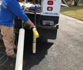 A worker in a blue shirt and khaki pants stands by a service truck labeled Emergency Well Pump Service, handling large pipes and equipment at the back of the vehicle on a paved surface outdoors. A worker in a blue shirt and khaki pants stands by a service truck labeled Emergency Well Pump Service, handling large pipes and equipment at the back of the vehicle on a paved surface outdoors.