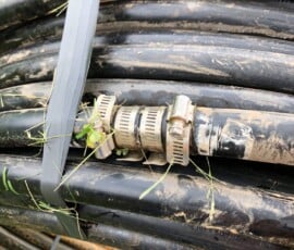A close-up of a bundle of black, dirt-streaked hoses secured with duct tape and metal hose clamps, lying on green grass with small blades and debris stuck to the surface. A close-up of a bundle of black, dirt-streaked hoses secured with duct tape and metal hose clamps, lying on green grass with small blades and debris stuck to the surface.