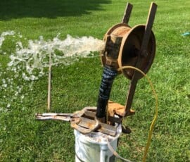 Water gushes forcefully from a pipe attached to a rusted pump mechanism on a grassy lawn, with houses and trees visible in the background. Water gushes forcefully from a pipe attached to a rusted pump mechanism on a grassy lawn, with houses and trees visible in the background.
