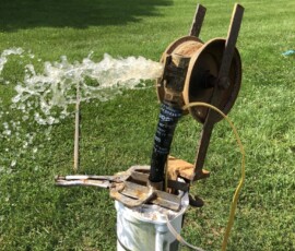 Water gushes forcefully from a pipe attached to a rusted pump mechanism on a grassy lawn, with houses and trees visible in the background. Water gushes forcefully from a pipe attached to a rusted pump mechanism on a grassy lawn, with houses and trees visible in the background.