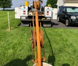 A rusty metal pipe with pulleys is being pulled from a white well casing in a grassy yard, with a utility truck and black SUV parked on a driveway in the background. Tools and equipment are visible on the truck bed. A rusty metal pipe with pulleys is being pulled from a white well casing in a grassy yard, with a utility truck and black SUV parked on a driveway in the background. Tools and equipment are visible on the truck bed.