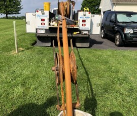 A rusty metal pipe with pulleys is being pulled from a white well casing in a grassy yard, with a utility truck and black SUV parked on a driveway in the background. Tools and equipment are visible on the truck bed. A rusty metal pipe with pulleys is being pulled from a white well casing in a grassy yard, with a utility truck and black SUV parked on a driveway in the background. Tools and equipment are visible on the truck bed.