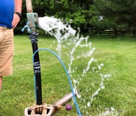 Water sprays out in an arc from a black pipe setup in a grassy yard. A person in shorts and a blue shirt stands nearby, partially visible. Trees and green grass are in the background. Water sprays out in an arc from a black pipe setup in a grassy yard. A person in shorts and a blue shirt stands nearby, partially visible. Trees and green grass are in the background.