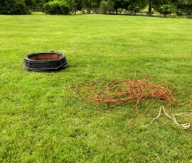 A coil of black tubing and a tangled bundle of orange and black cables lie on a grassy lawn with trees and bushes in the background under a cloudy sky. A coil of black tubing and a tangled bundle of orange and black cables lie on a grassy lawn with trees and bushes in the background under a cloudy sky.