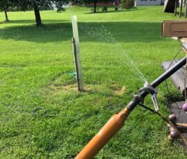 A homemade sprinkler made from a yellow pipe sprays water across a green lawn on a sunny day, with houses, trees, and a wooden post visible in the background. A homemade sprinkler made from a yellow pipe sprays water across a green lawn on a sunny day, with houses, trees, and a wooden post visible in the background.